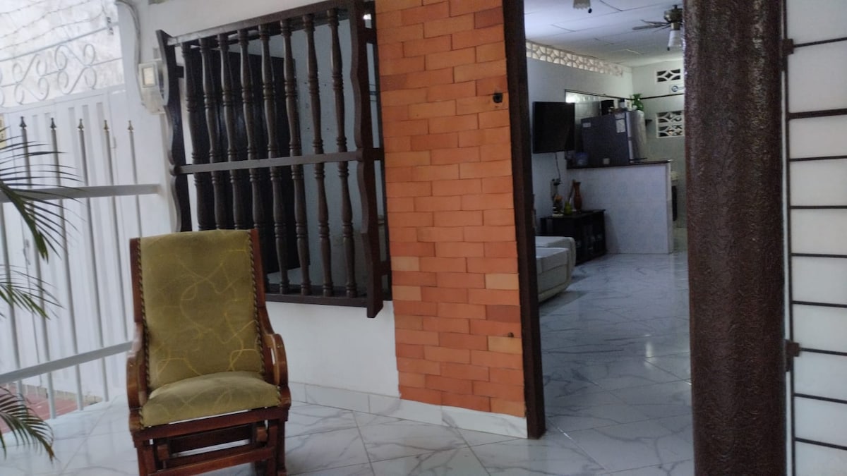 A welcoming entrance area features a wooden chair positioned on a polished marble floor. Walls are adorned with a mix of brick and white finishes. A glimpse of a living space is visible in the background, showcasing minimalist decor and modern furnishings.