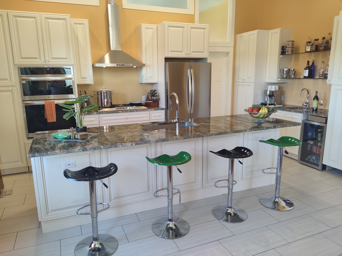 A spacious kitchen features white cabinetry, a large granite island with four bar stools, and modern appliances. A stainless steel range hood and a double oven are visible, along with a refrigerator. Light-colored tiles enhance the kitchen's bright and clean atmosphere, with decorative shelving displayed against the wall.