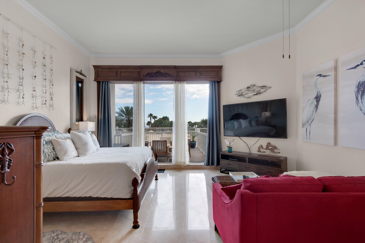 A spacious studio apartment features a king bed with white bedding, a vibrant red couch, and a large flat-screen TV mounted on the wall. Natural light fills the room through glass doors that open to a patio, complementing the light-colored walls and marble flooring.