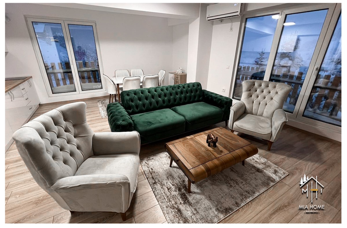 A spacious living area features a green tufted sofa, two light-colored armchairs, and a wooden coffee table. Large windows offer views of the outside, allowing natural light to brighten the room. A nearby dining table is visible, accommodating six guests comfortably.
