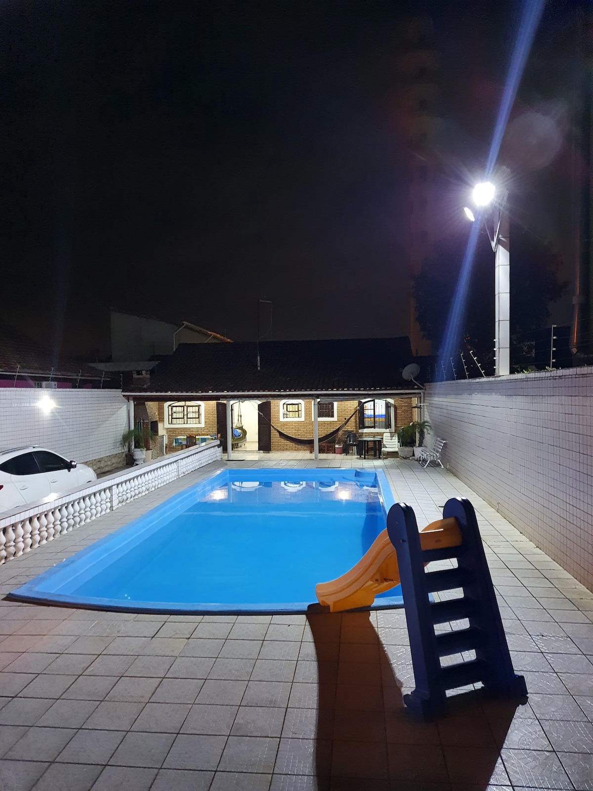 A well-lit outdoor area features a large blue swimming pool, complemented by a small slide on the edge. Surrounding spaces include a house in the background, with white tiles paving the area and comfortable seating arrangements.