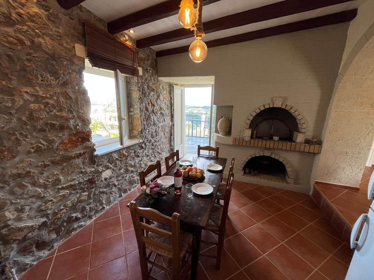 A rustic dining area features a wooden table set with plates, glasses, and a centerpiece of fruit. Exposed stone walls and wooden beams create a warm atmosphere. A traditional oven is visible, and large windows offer views of the landscape.
