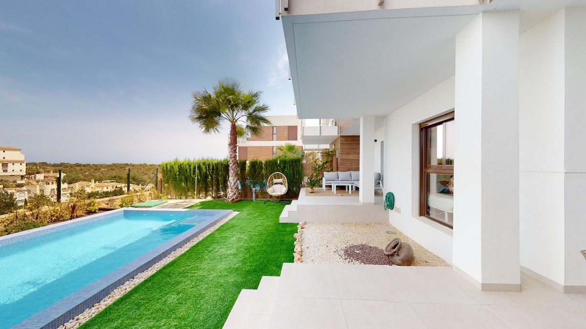 The image captures a private pool surrounded by a manicured garden. A comfortable seating area is visible next to the pool, alongside a shaded terrace. Lush greenery and distant views of the countryside complement the tranquil setting.