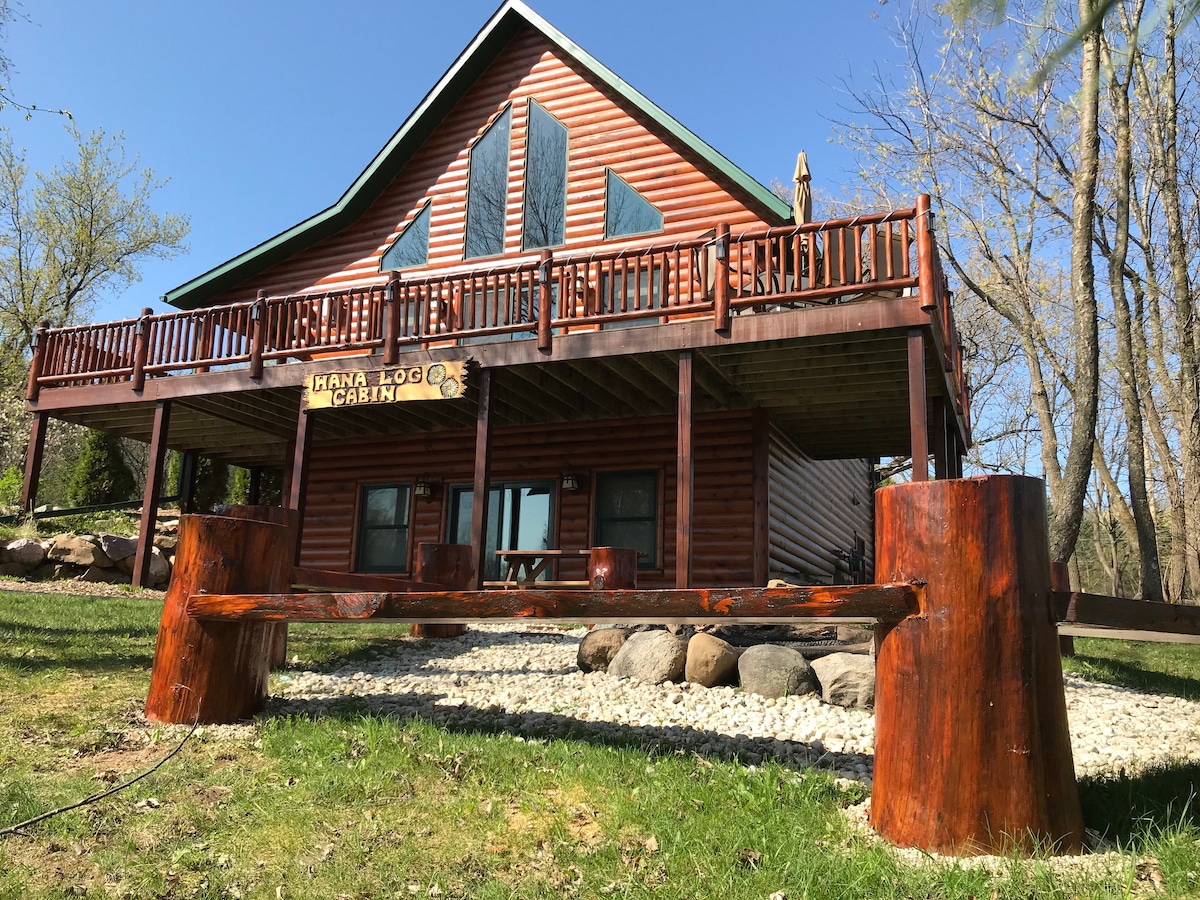 A rustic log cabin is set against a backdrop of greenery. The expansive wrap-around deck features wooden rails and overlooks a gravel area with large stones. Two rounded wooden benches are positioned in front, enhancing the cabin's natural aesthetic.