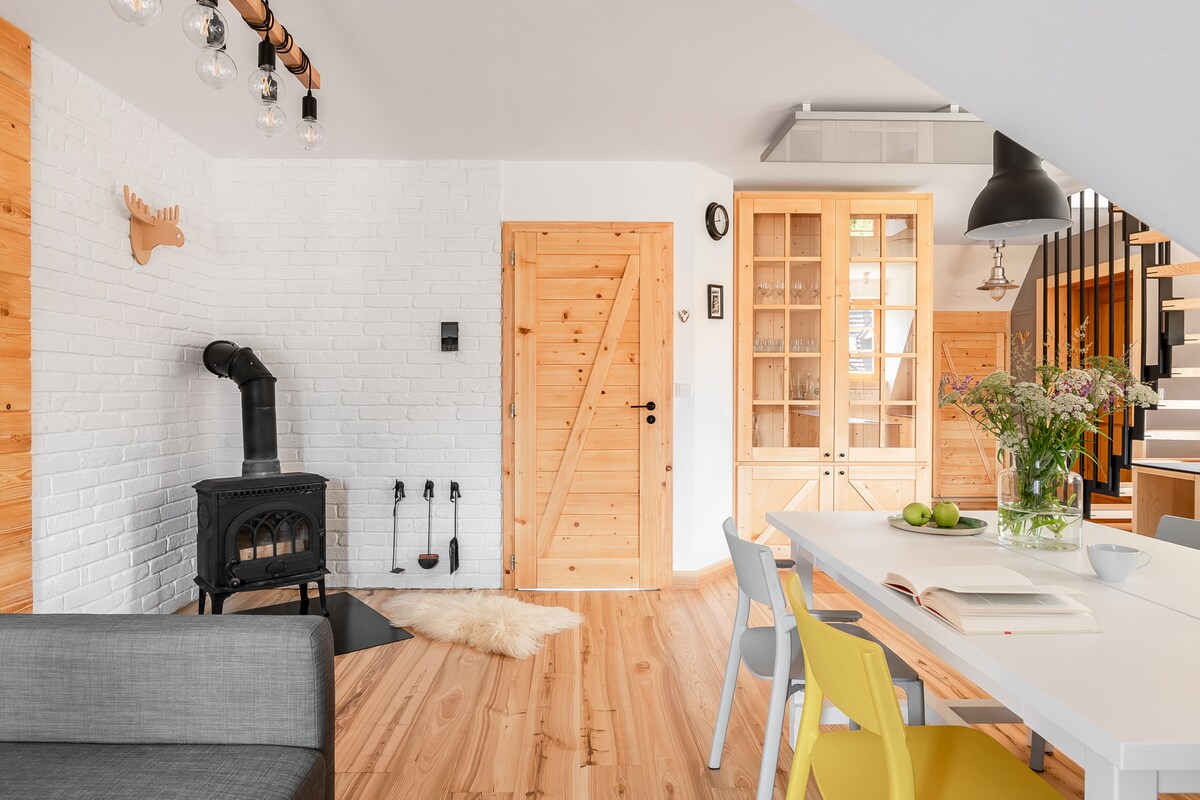 A charming living space is presented, featuring a wood-burning stove against a white brick wall. A light wood dining table is surrounded by colorful chairs. Natural light fills the area, highlighting wooden flooring and a cozy texture of a decorative rug.