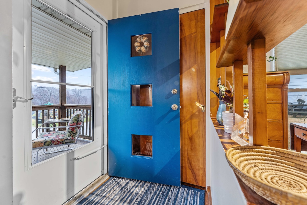 A bright blue front door features three decorative cut-out windows and opens into a welcoming entryway. A patterned area rug covers the hardwood floor, with a glimpse of a cozy chair visible through the adjacent glass door that leads to a porch.