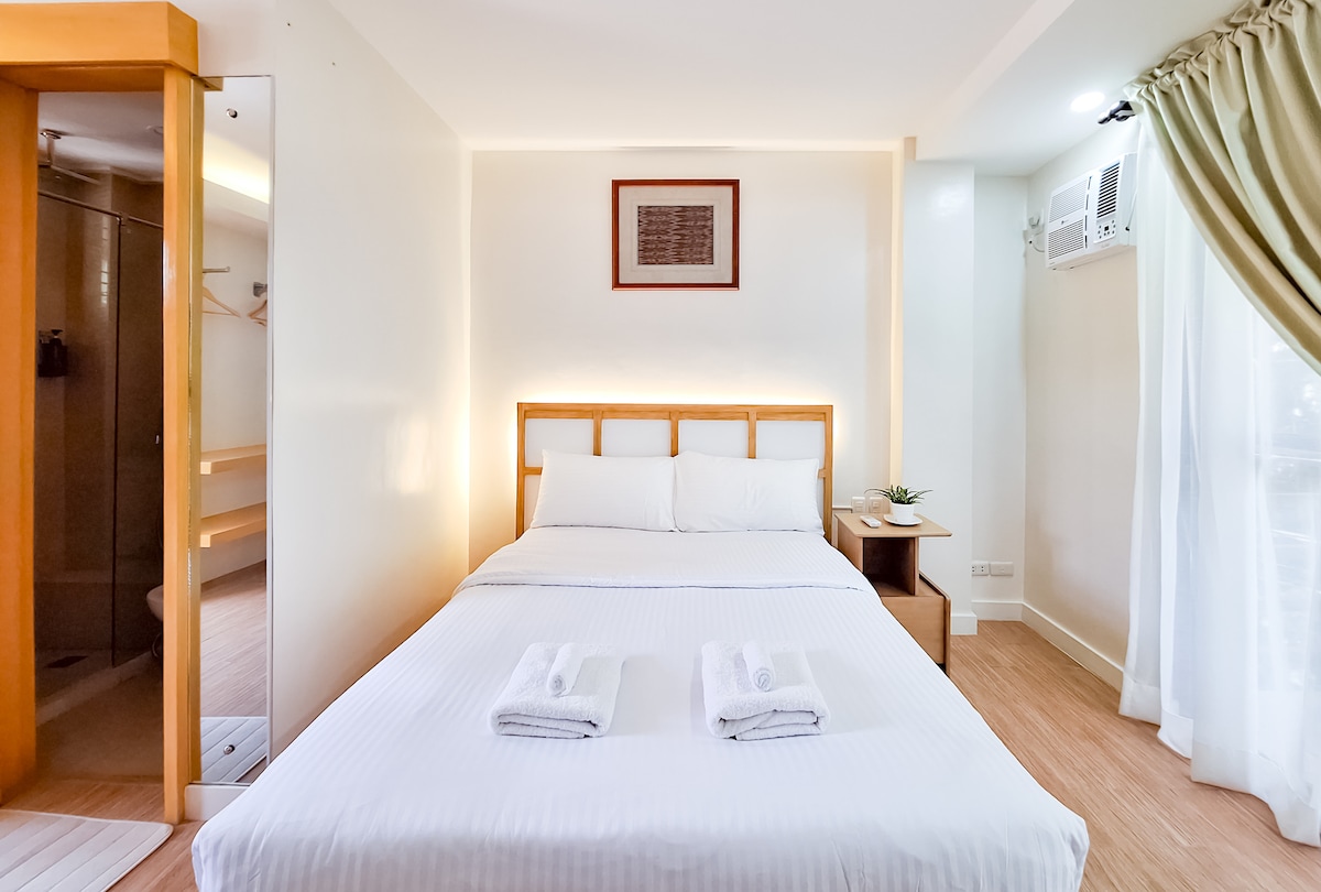 A well-lit bedroom features a spacious bed with neatly arranged white linens and couple of white towels atop. A small decorative plant sits on the bedside table, while natural light enters through a nearby window framed by soft drapes. The en-suite bathroom is visible to the left.
