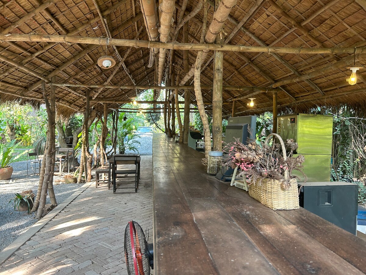 An open-air communal dining space is framed by bamboo and thatched roofing, featuring a long wooden table. Lush greenery surrounds the area, with soft lighting illuminating the path. A basket filled with dried plants rests on the table, adding a rustic touch to the inviting atmosphere.