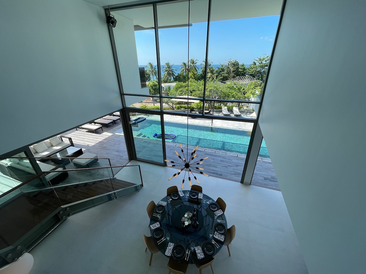 A spacious dining area is visible, featuring a round table set for guests. Floor-to-ceiling windows provide a view of the outdoor pool surrounded by lounge chairs and lush greenery, with the Andaman Sea sparkling in the background.