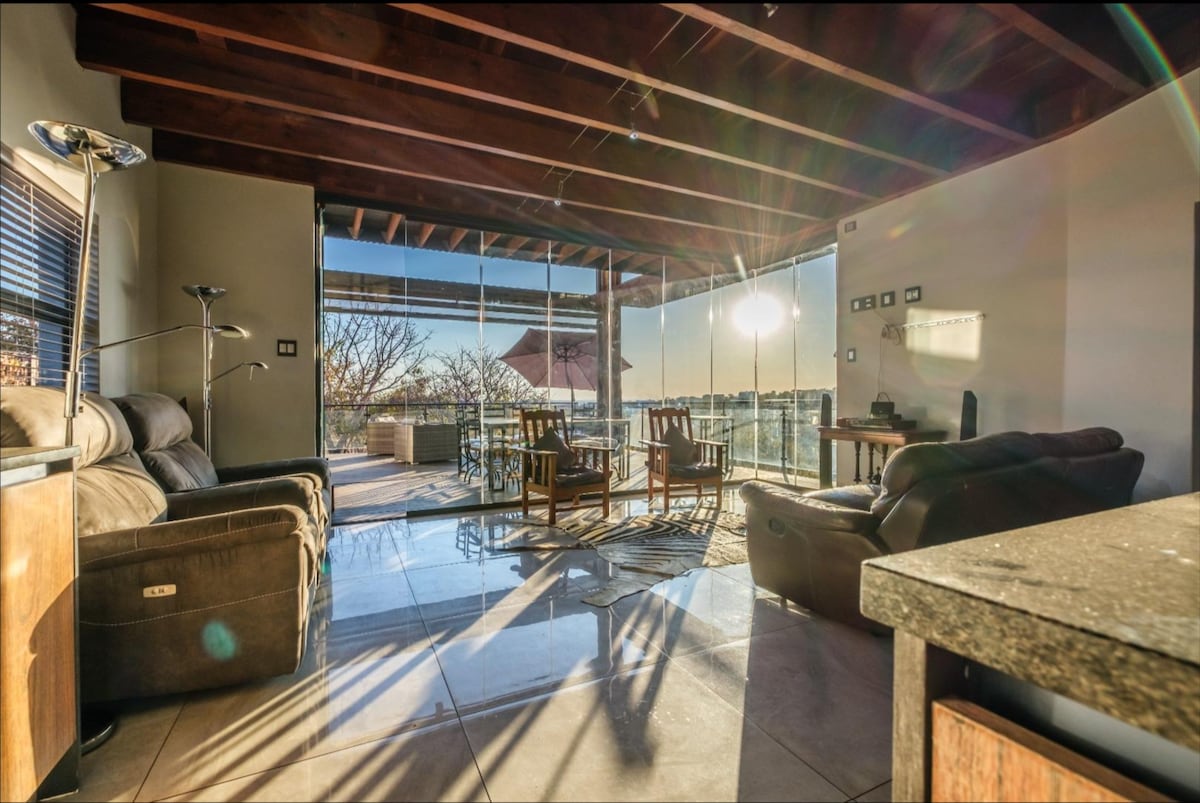 A spacious living area is highlighted by large glass doors that invite natural light, showcasing a serene patio view. Comfortable seating is arranged for relaxation, with a modern lamp and a sleek countertop visible, enhancing the welcoming ambiance of the space.