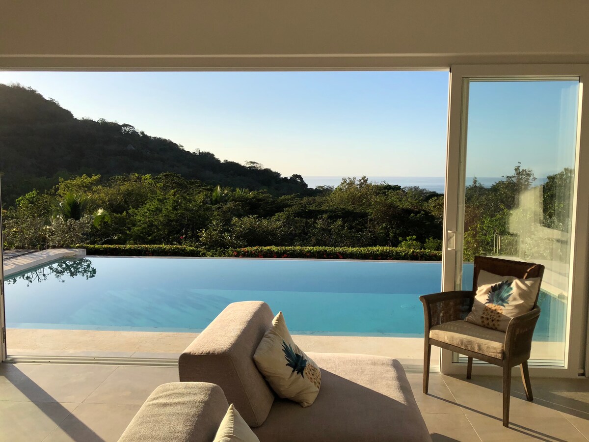 A spacious living area featuring large glass doors that open to a stunning view of lush greenery and the ocean beyond. A comfortable sofa and a wicker chair offer relaxation options, while the serene infinity pool glistens under natural light.