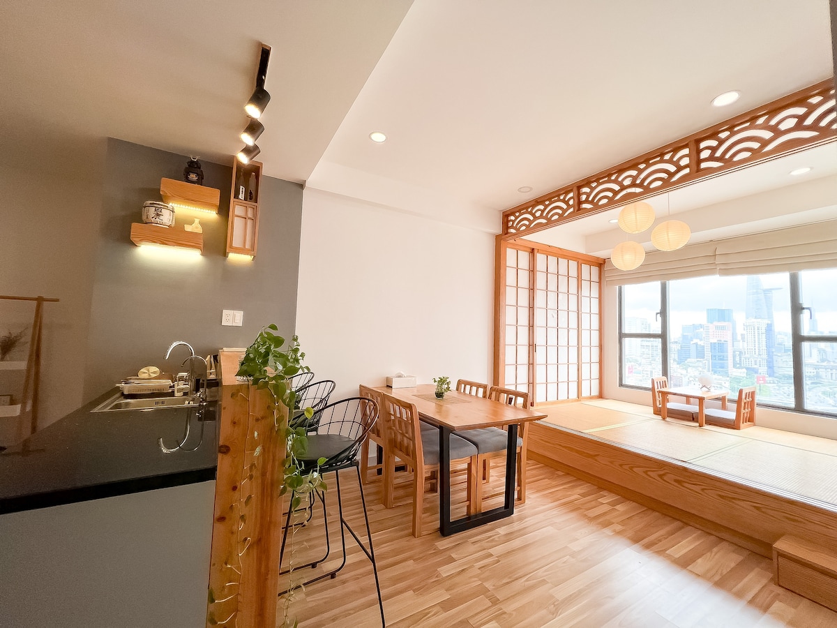 A modern kitchen area features sleek black countertops and wooden accents, complemented by minimalistic shelving. Open seating is provided by a dining table with black chairs, adjacent to a cozy nook with tatami mats and a wall of large windows offering city views.