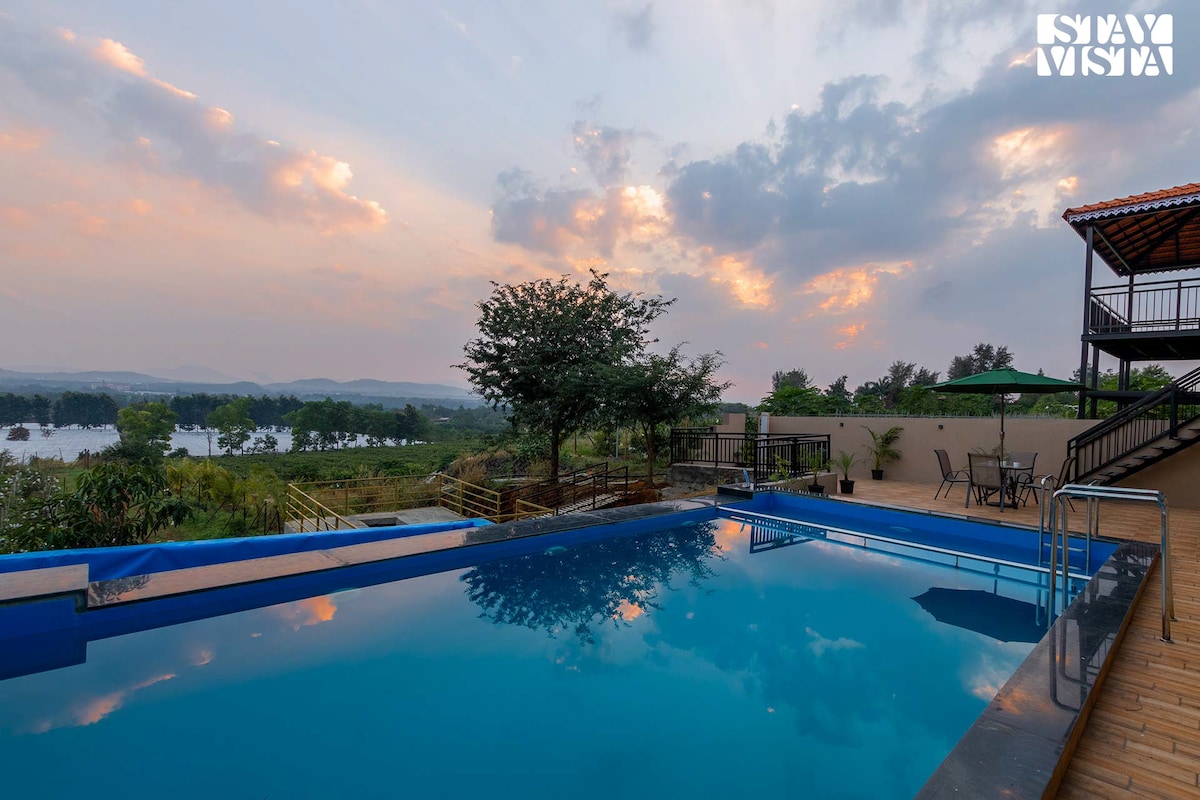 A serene outdoor pool reflects the colors of the evening sky, surrounded by lush greenery. An alfresco dining area is visible near the pool, with comfortable seating and umbrellas for shade. In the background, expansive views of the farms are framed by the landscape.