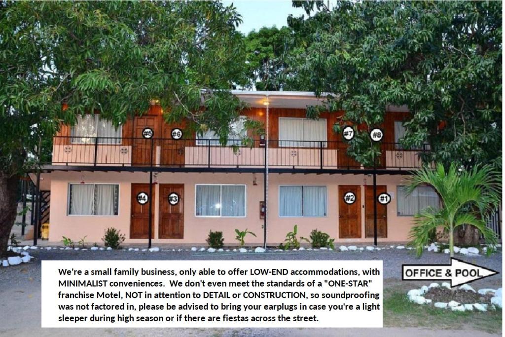 A two-story motel building is depicted, surrounded by lush greenery. Each room features a private balcony with white railings. The pool area is visible in the foreground, offering a relaxing outdoor space. The exterior is painted in soft hues, contributing to a tranquil ambiance.