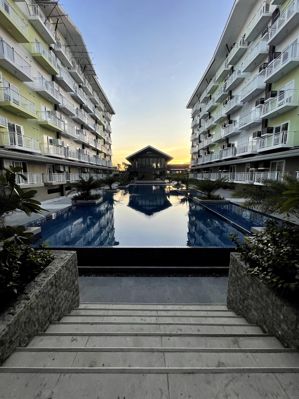 Two modern condo buildings frame a serene pool area, reflecting the colors of a sunset. Lush greenery and palms surround the pool, creating a tranquil environment. The scene is peaceful, emphasizing the inviting natural light and calm waters.