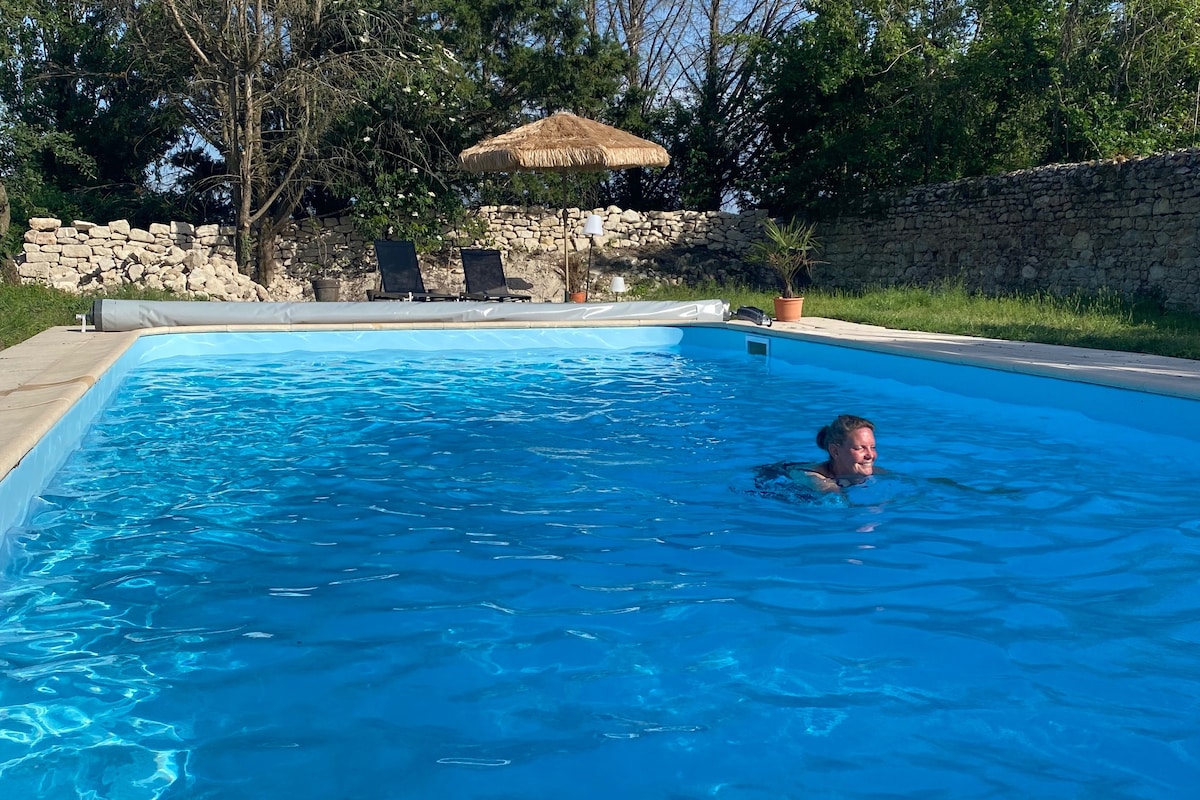 A clear, inviting outdoor pool is set within a grassy area, surrounded by trees and stone walls. Sun loungers are positioned adjacent to the pool, and a shaded umbrella provides a relaxing spot nearby.