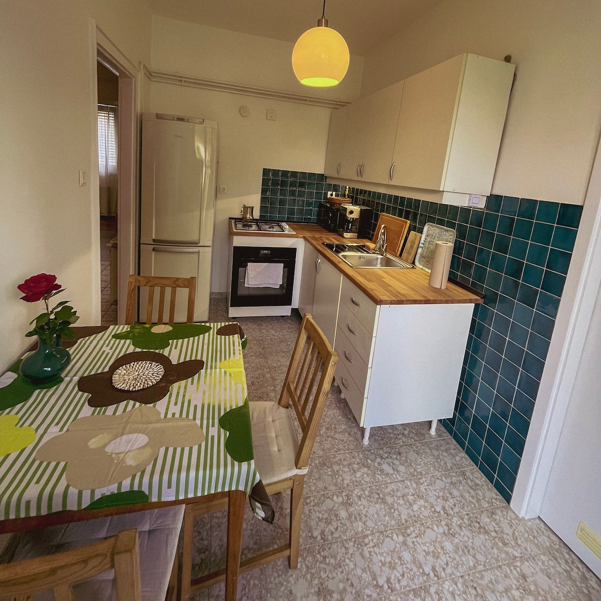 The kitchen displays a blend of vintage and modern elements, featuring a wooden countertop and turquoise tiled backsplash. A dining table with an artistic tablecloth is set for four, with a vase of red roses adding a touch of color. Appliances include a gas stove and a refrigerator.