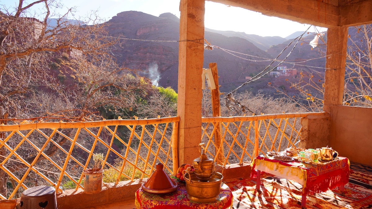 A terrace offers a sweeping view of the surrounding mountains and valleys. A low table adorned with traditional Moroccan dishes sits on a vibrantly colored cloth. Sunlight filters through the wooden railing, illuminating the rustic architecture and natural elements of the landscape.