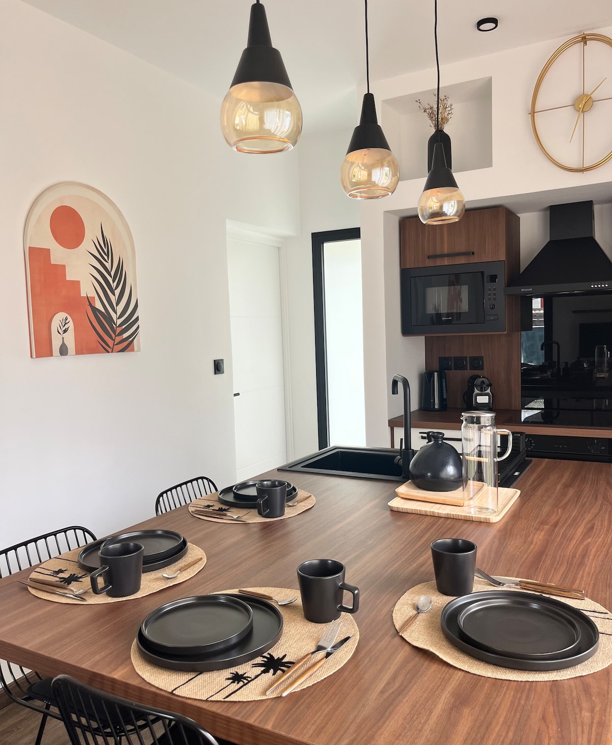 A dining area is presented with a wooden table set for four. Black plates and mugs are arranged on woven placemats. Modern pendant lights hang from the ceiling above the table, illuminating a wall art piece that features warm colors and a tropical motif.