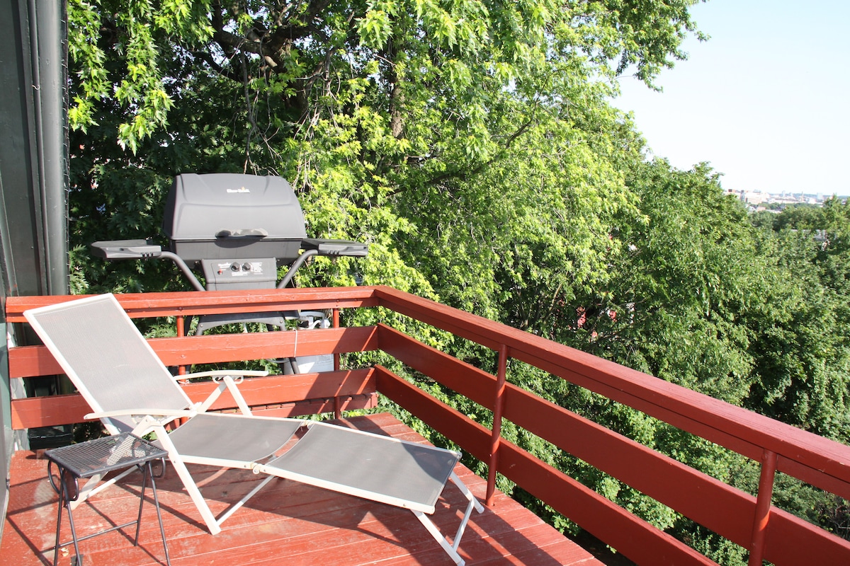 An outdoor balcony features a red wooden railing overlooking lush greenery. A grill is placed on one side, while two lounge chairs are positioned for relaxation. Sunlight filters through the trees, providing a serene atmosphere.