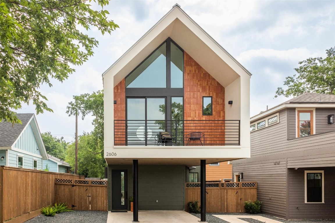 The exterior of a modern home is presented, featuring a distinctive triangular roofline and large windows that allow for ample natural light. A covered patio area with two chairs is visible on the balcony. Surrounding greenery and wooden fencing contribute to the overall design.