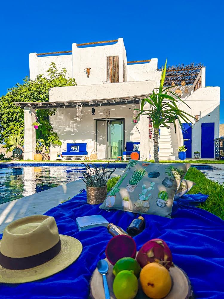 A serene outdoor setting presents a poolside view of a white-stucco house, accentuated by vibrant blue accents. A picnic setup features a blue blanket adorned with a pillow and fresh fruits, complemented by a sun hat resting nearby, inviting relaxation in the tranquil environment.