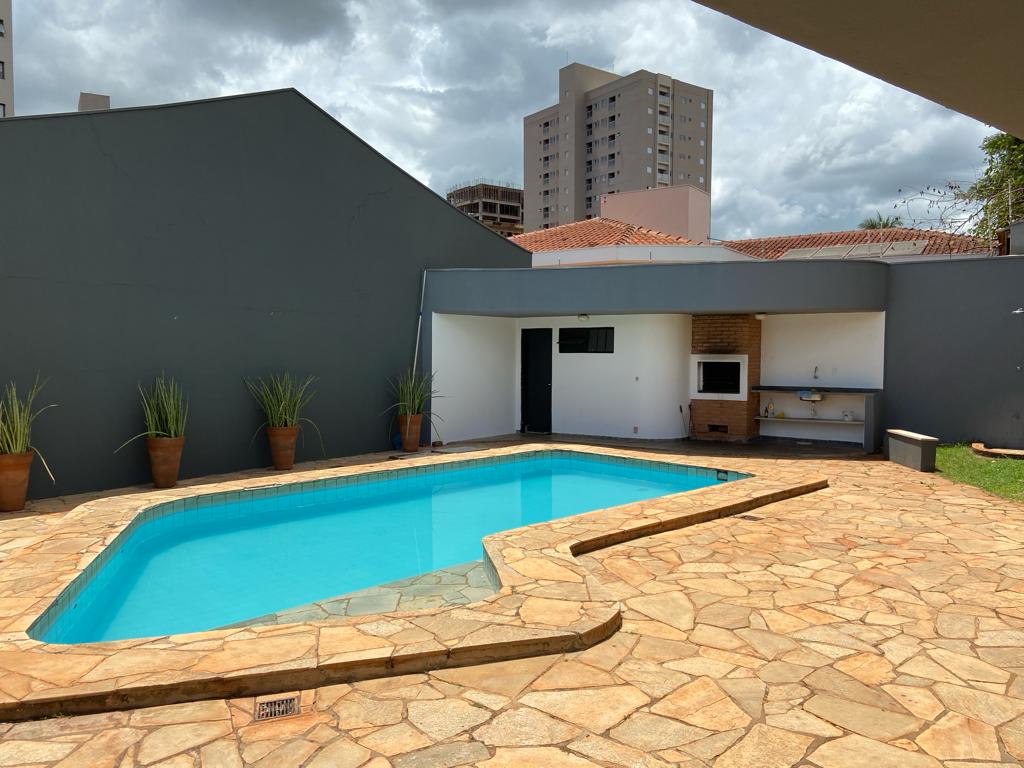 An outdoor area features a bright pool surrounded by a stone deck. Potted plants line the space, and a barbecue area is visible with a television mounted on the wall. The surroundings includes a neutral grey wall and a view of buildings in the background.