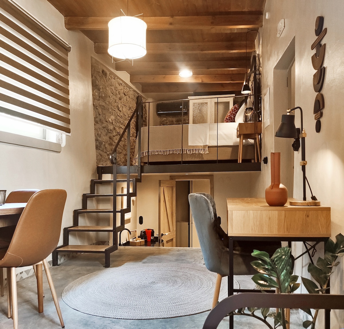 An open loft suite is showcased, featuring a double bed elevated on a wooden platform. A modern kitchenette occupies one side, while a round rug centers a seating area with a chair and dining table. A wooden staircase leads to the sleeping space, illuminated by soft lighting.