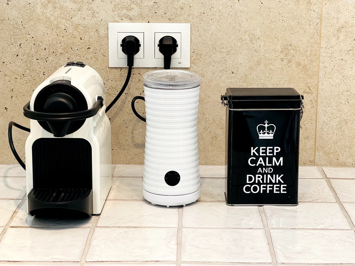 A coffee station is displayed on a tiled surface, featuring a compact espresso machine, a white coffee grinder, and a black storage container. The storage container bears a motivational phrase about coffee. Electrical outlets are visible in the background.