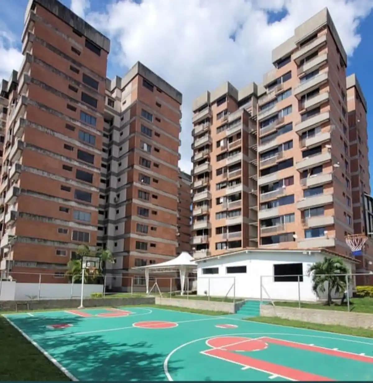 Two large residential buildings are positioned adjacent to a basketball court, featuring vibrant green surfaces marked with red and white lines. The buildings boast ample balconies and windows, surrounded by well-maintained landscaping, creating an inviting outdoor space.