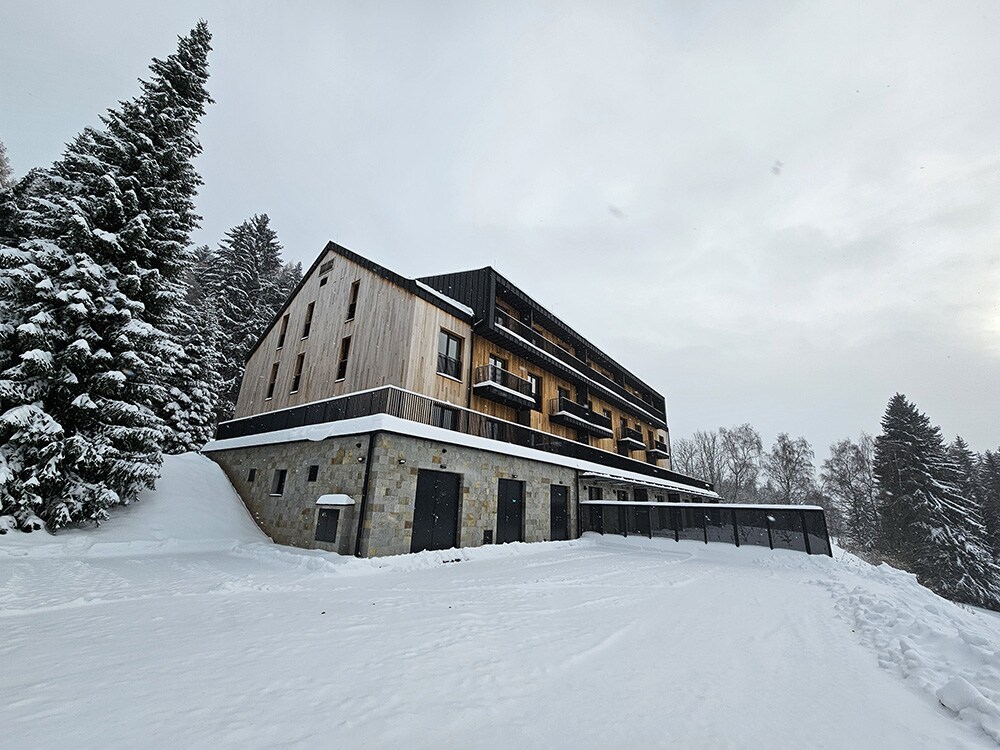 A modern building is set against a snowy landscape, showcasing a combination of wood and stone exterior. Balconies are featured on the upper floors, while the ground level displays a structured entrance. Snow blankets the surrounding area, enhancing the serene winter atmosphere.
