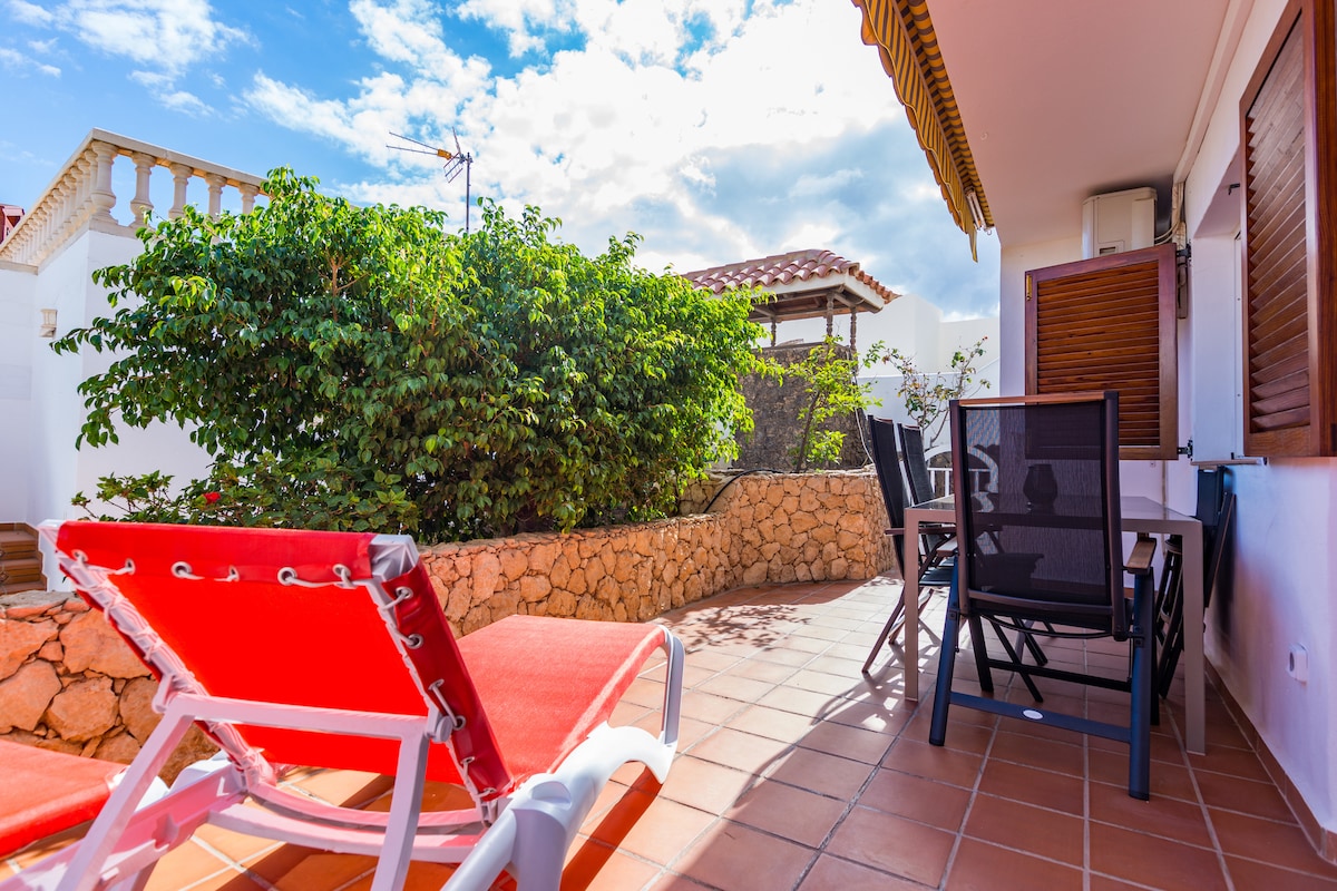 An outdoor terrace features sun loungers positioned beside a stone wall adorned with lush greenery. A dining area with chairs is visible, creating a space for outdoor relaxation. The terrace is partially shaded, providing a comfortable atmosphere for enjoying the outdoors.