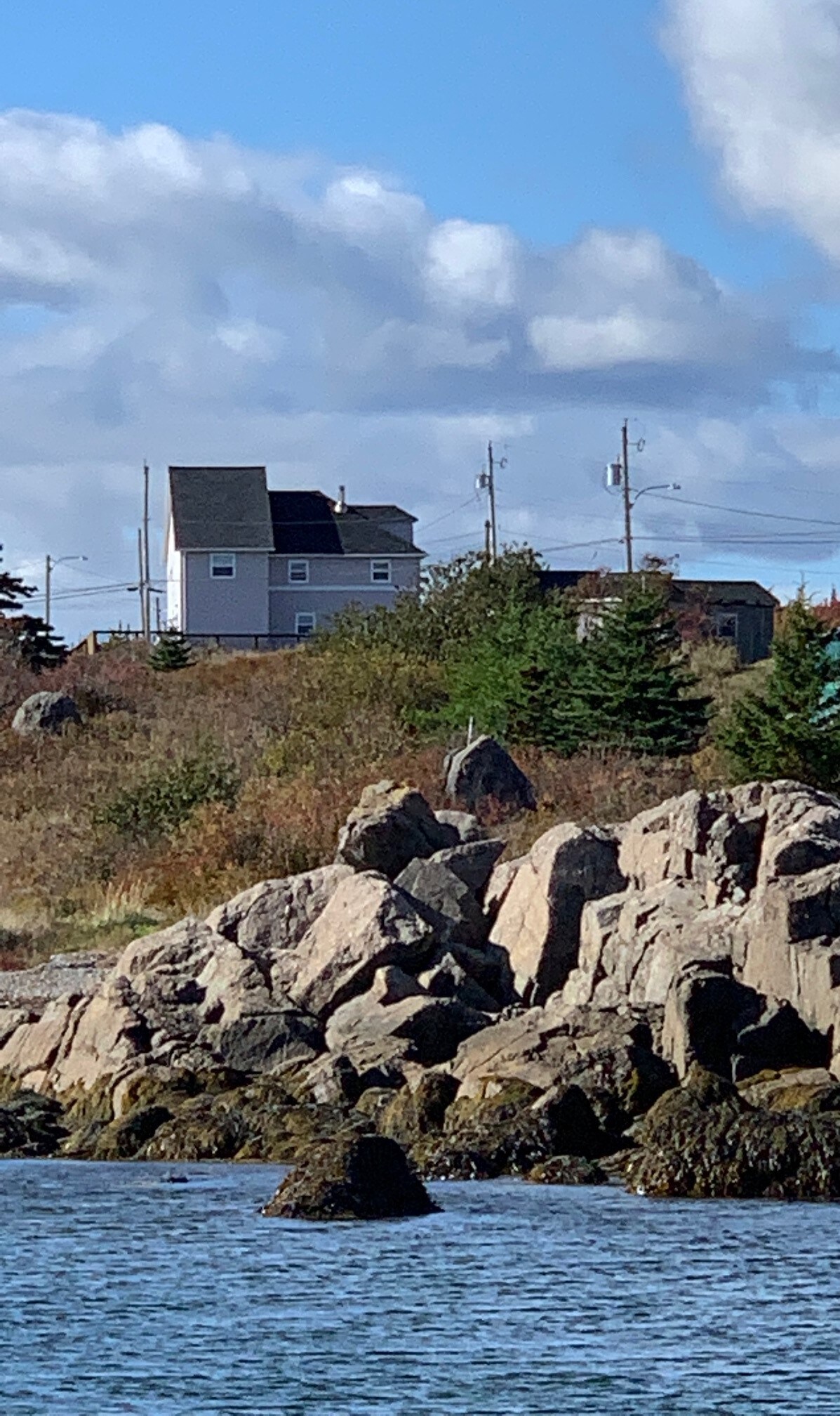 A two-story house is positioned on a grassy hill, surrounded by rocky formations and coastal vegetation. The structure features a dark roof and light siding, contrasting with the blue sky and scattered clouds above. The scene captures a serene coastal landscape.