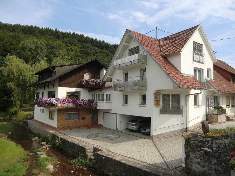The exterior of a well-maintained multi-level house is shown, featuring a blend of white and dark wood finishes. Vibrant flower boxes adorn the balconies. A spacious driveway is visible, accommodating parked vehicles. Lush greenery from the surrounding landscape enhances the peaceful setting.