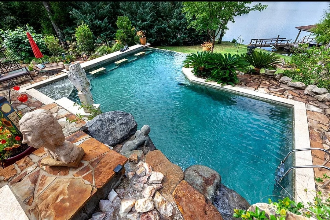 An inviting infinity pool is surrounded by natural stone and vibrant landscaping. The pool's clear water reflects the green of nearby trees, with a lounging area featuring a red umbrella visible in the background, enhancing the serene outdoor setting.