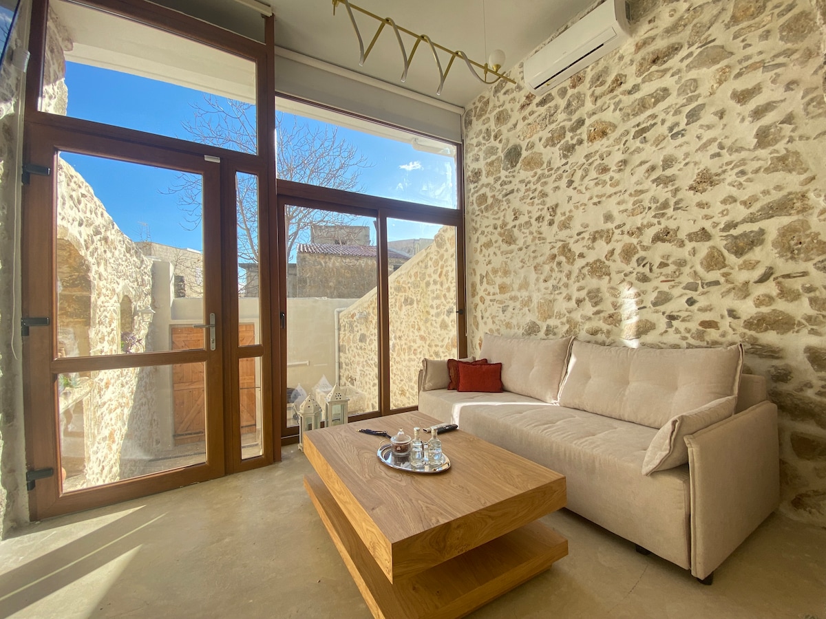 A cozy living area features a light-colored, plush sofa complemented by a warm wooden coffee table. Natural light floods in through expansive glass doors, which open up to a charming outdoor space surrounded by traditional stone walls.