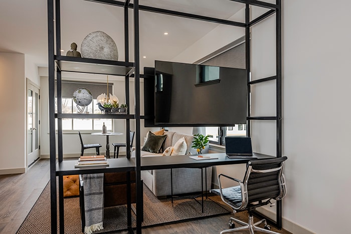 A modern workspace is featured with a sleek black desk and an ergonomic chair. A large flat-screen television is mounted above the desk, and a cozy seating area is visible in the background. Natural light filters through the windows, illuminating the contemporary decor.