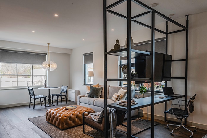 A modern living area features a spacious sofa and an ottoman, complemented by a sleek work desk and a cozy dining table for two. Ample natural light filters through large windows, enhancing the inviting atmosphere of this thoughtfully designed space.
