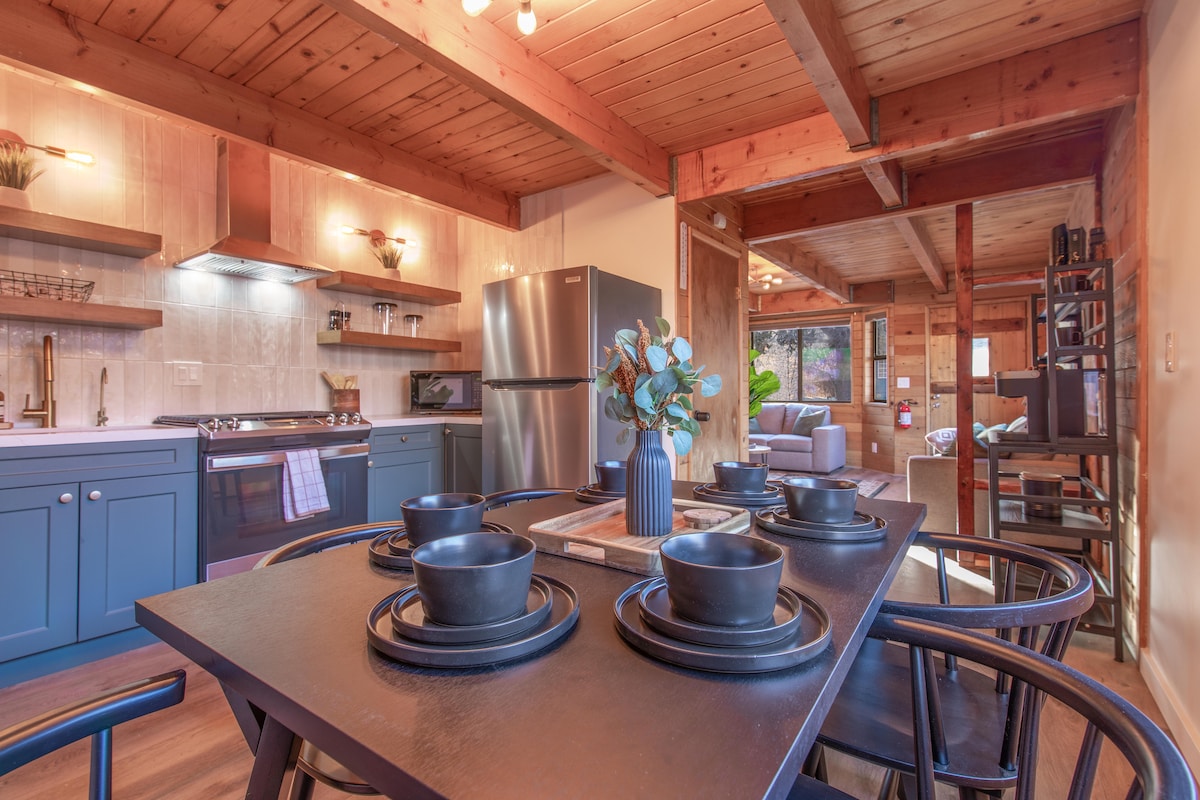 A modern kitchen is presented with wooden beams and warm tones. A dining table is set with black bowls and a central vase, while stainless steel appliances and blue cabinetry create a functional cooking space. Soft lighting enhances the welcoming ambiance.