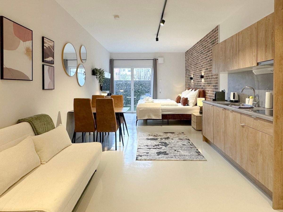 A spacious living area features a comfortable sofa and dining table. The arrangement includes a cozy sleeping area with a double bed and soft bedding. Natural light fills the room, highlighting the modern kitchen with wooden cabinetry and essential appliances. Large windows provide views of the private garden.