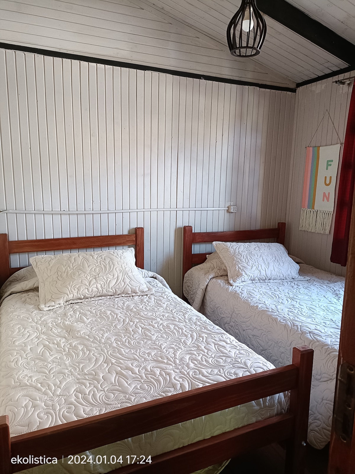 A cozy bedroom is presented, featuring two wooden beds covered with light-colored bedding. The walls are adorned with vertical white paneling, and a decorative wall hanging adds a touch of color. A pendant light is suspended from the ceiling, contributing to the room's ambiance.