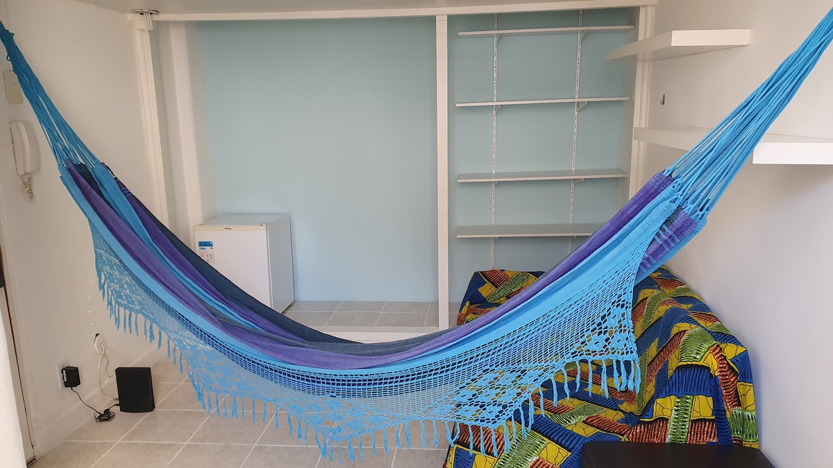 A vibrant blue hammock is suspended in a well-lit space, inviting relaxation. The room features a cozy patterned couch in bright colors. A small white refrigerator sits against the wall, and shelving provides additional storage options.