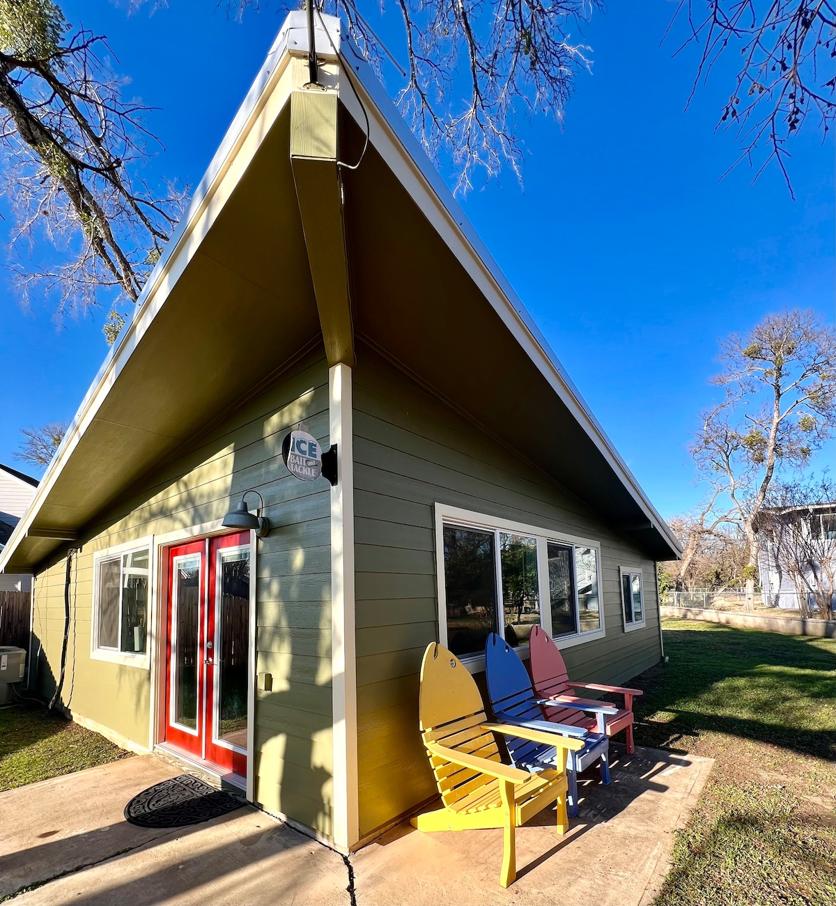 A vibrant green cabin exterior is showcased, featuring a slanted roof and large windows. Colorful Adirondack chairs in blue, yellow, and red are positioned along the side, inviting relaxation. A clear blue sky forms a bright backdrop, enhancing the outdoor appeal.