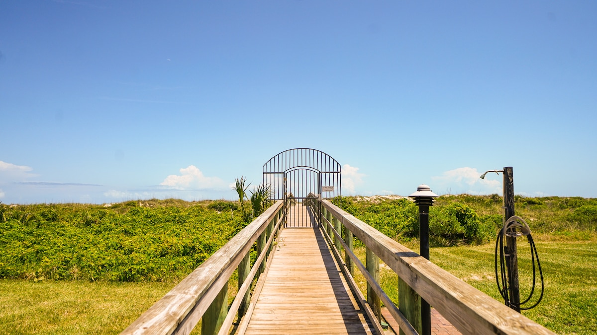 A wooden boardwalk leads through lush greenery towards a gated entrance, with the ocean visible in the distance. Clear blue skies and a few clouds create a serene backdrop, inviting guests to explore the beach beyond.