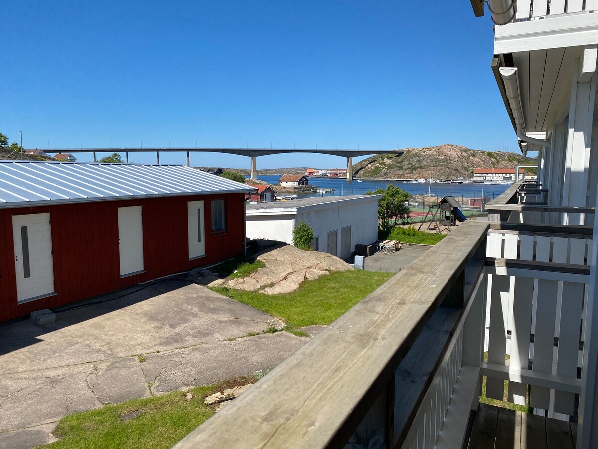 A view from the balcony showcases a bridge spanning across the water, with rocky terrain and grassy areas below. Red buildings and a paved area are visible in the foreground, providing a glimpse of the surrounding landscape. Clear blue skies enhance the scenery.