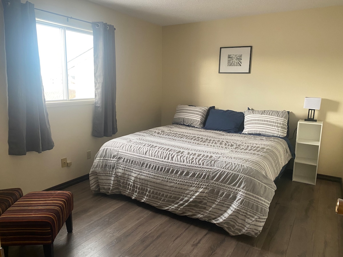 A bright sleeping area features a spacious bed with a striped comforter. Natural light enters through a nearby window, complemented by light curtains. A small bedside lamp and a white shelving unit are situated next to the bed, alongside a striped upholstered chair.