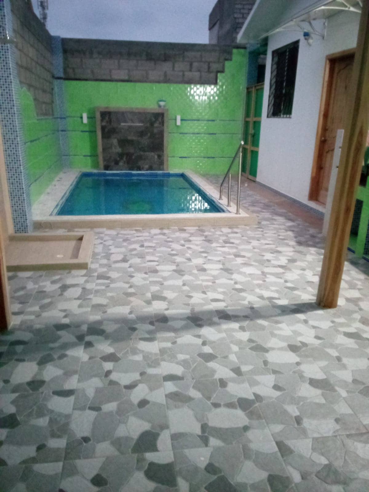 An outdoor area features a small pool surrounded by vibrant green tiles. Smooth, textured flooring extends toward the pool, enhancing the space's inviting atmosphere. A doorway leads into an adjoining area, framed by wooden columns that provide structural support.