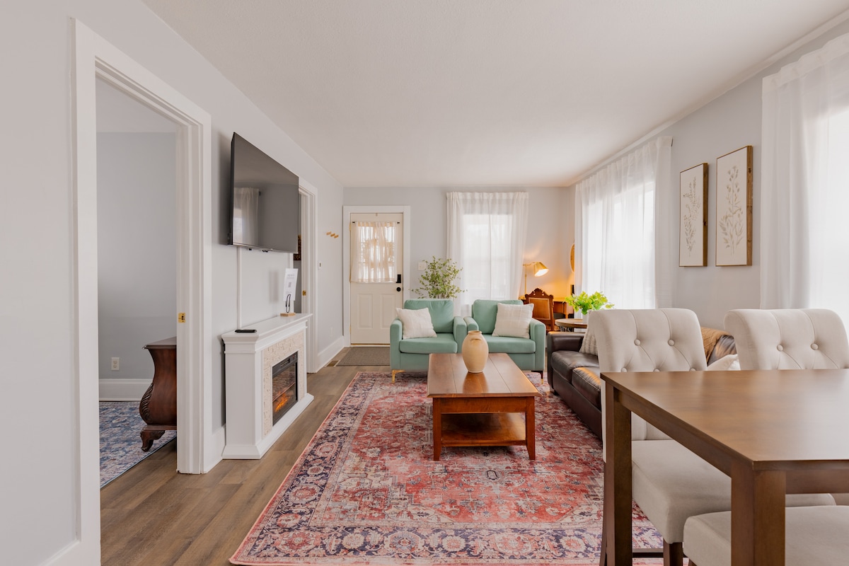 A welcoming living area features a mix of light and dark furnishings, including a plush seating arrangement with a coffee table at the center. Natural light floods through sheer curtains, highlighting decorative art on the walls and a television mounted above a fireplace.