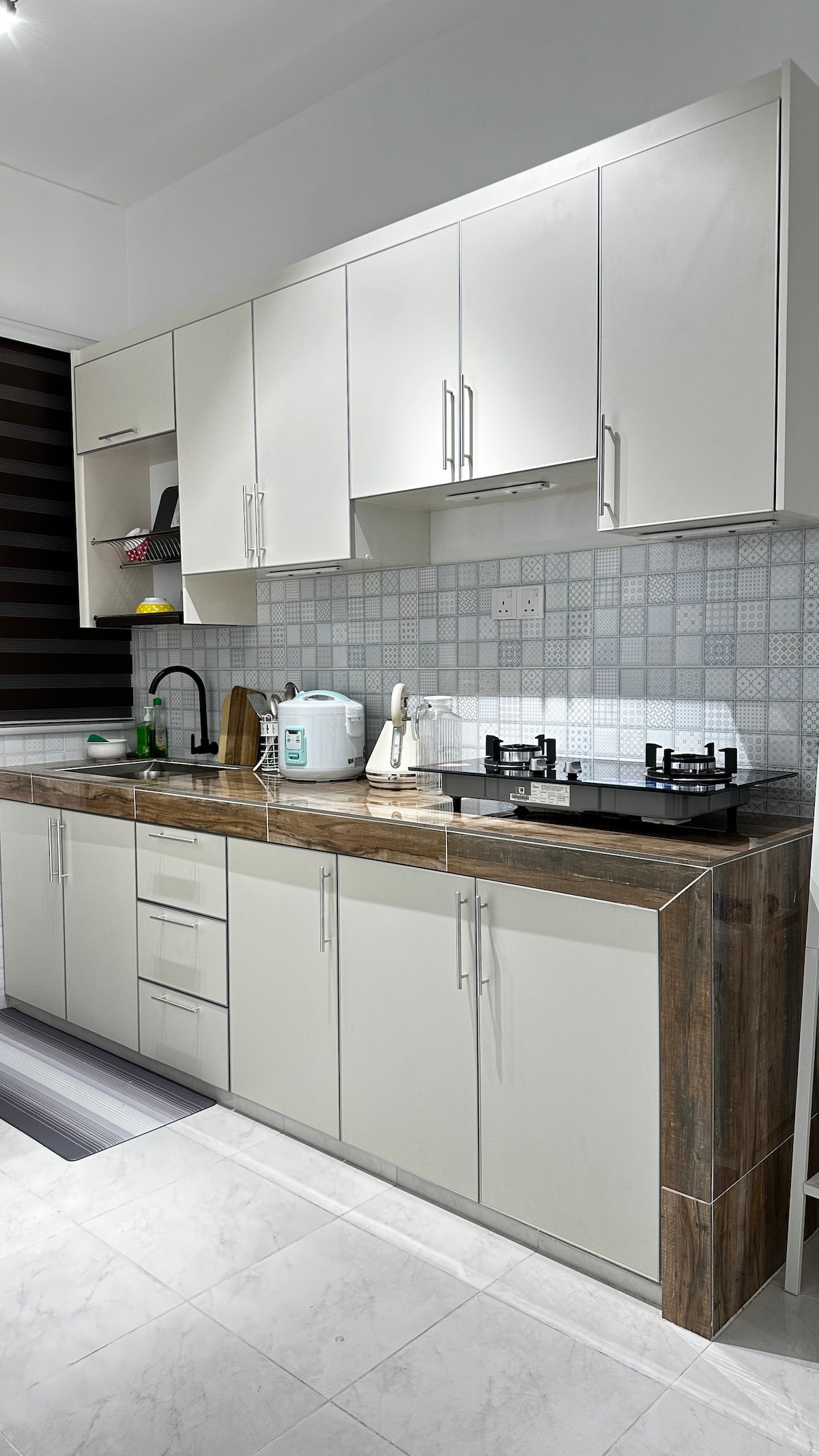 A modern kitchen is presented with sleek white cabinetry and a wooden countertop. The space features a gas stove with two burners, a kettle, and various kitchen essentials. A textured backsplash adds interest, while the flooring displays a combination of light tiles.