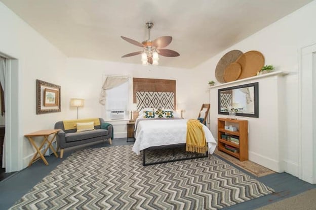 The spacious bedroom features a queen bed with a light-colored blanket and decorative pillows. A small seating area with a grey couch is present. A chevron-patterned rug covers the floor, while stylish wall decor and a ceiling fan add character to the bright, welcoming space.